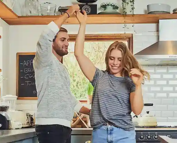 A man and woman smile and dance together in a modern kitchen, surrounded by cooking appliances and utensils.